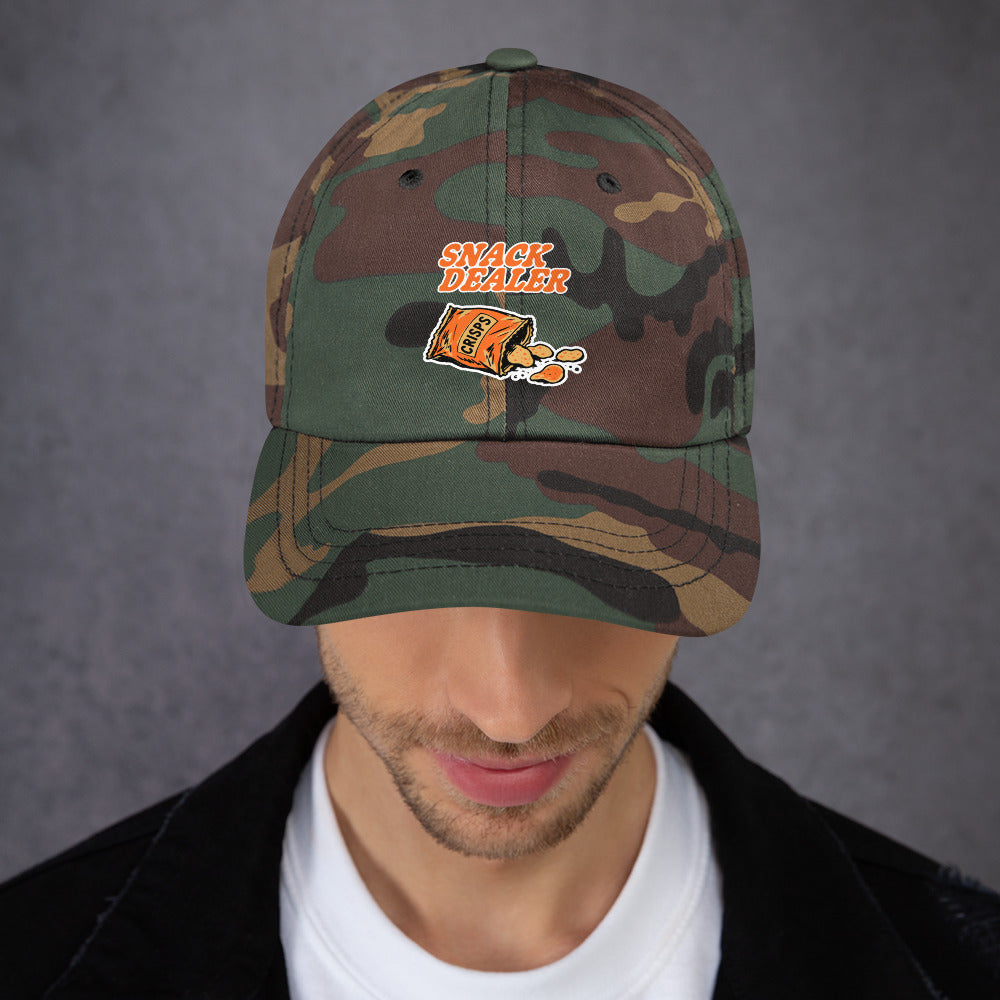 A man wearing a camouflage baseball cap with a custom embroidered patch that reads 'SNACK DEALER' and shows a cartoon character. The man is looking down, and the background is a textured gray wall.