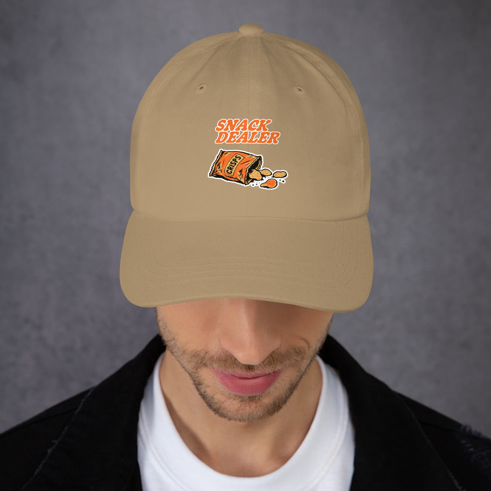 A man wearing a beige baseball cap with an embroidered graphic that reads 'SNACK DEALER' above an orange bag illustration, photographed against a gray textured background.