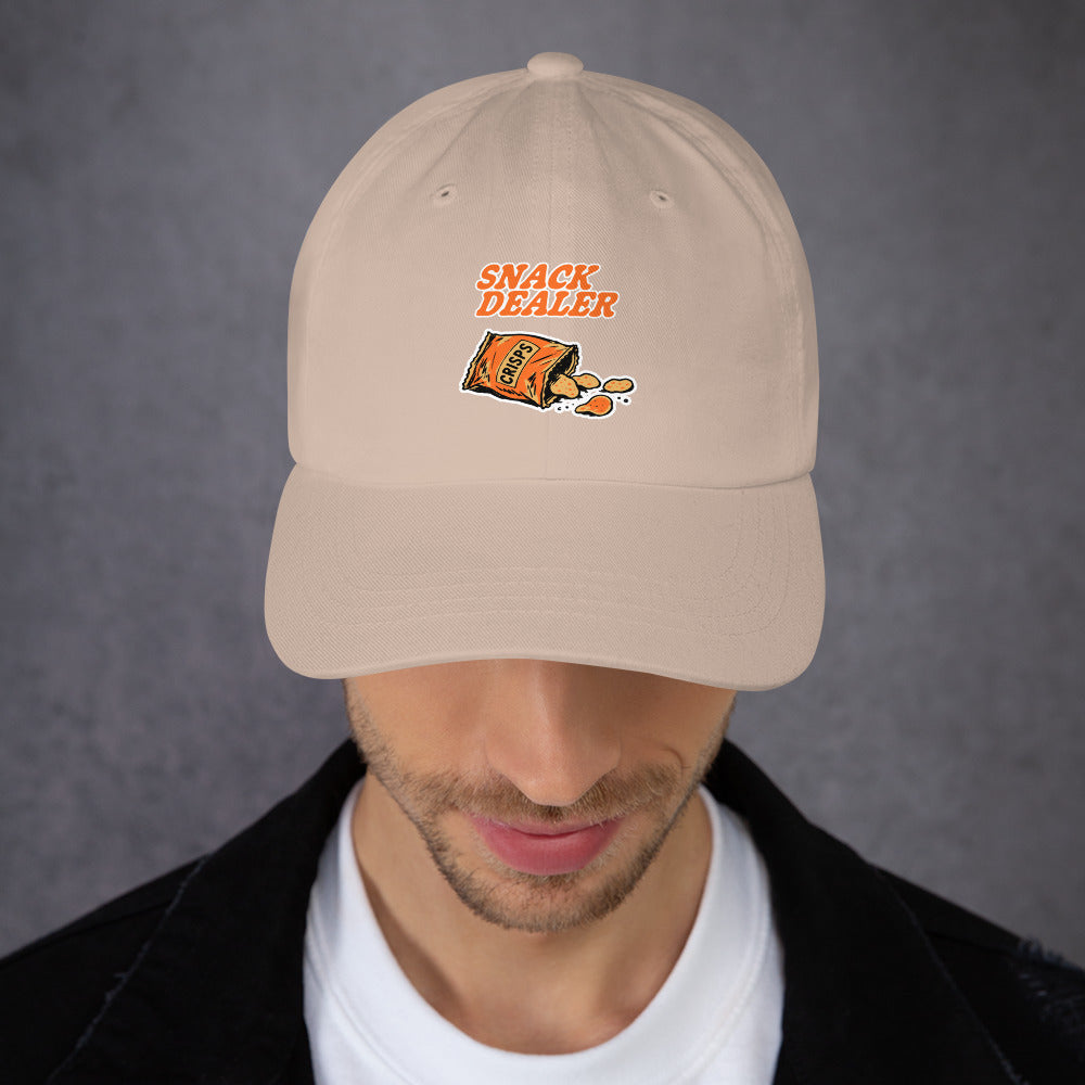 A man wearing a beige baseball cap with an embroidered graphic that reads 'SNACK DEALER' above an illustration of an open snack bag spilling chips. The man is looking down, and the background is a textured gray wall.
