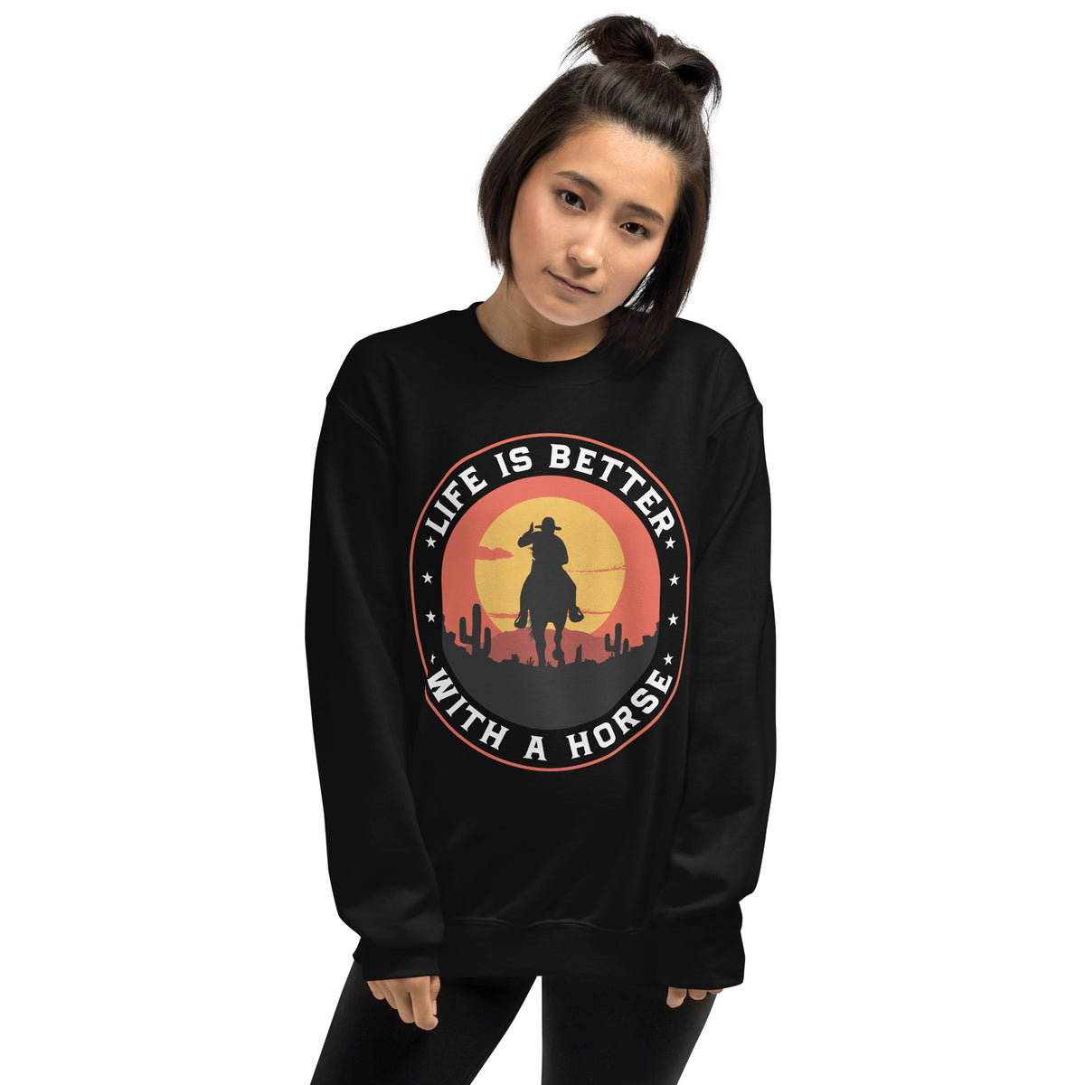 A woman wearing a black crewneck sweatshirt with a circular graphic on the chest that reads 'LIFE IS BETTER WITH A HORSE' around a cowboy silhouette against a sunset.
