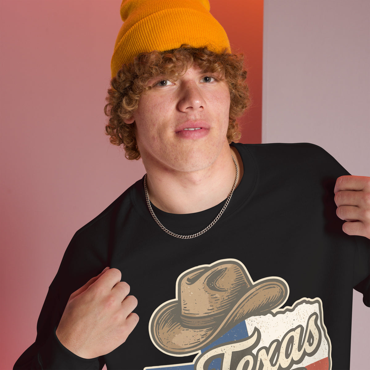 A young man wearing a bright yellow beanie and a black crewneck sweatshirt with a graphic of a cowboy hat and the word 'Texas' on the front, posing against a split pink and orange background.