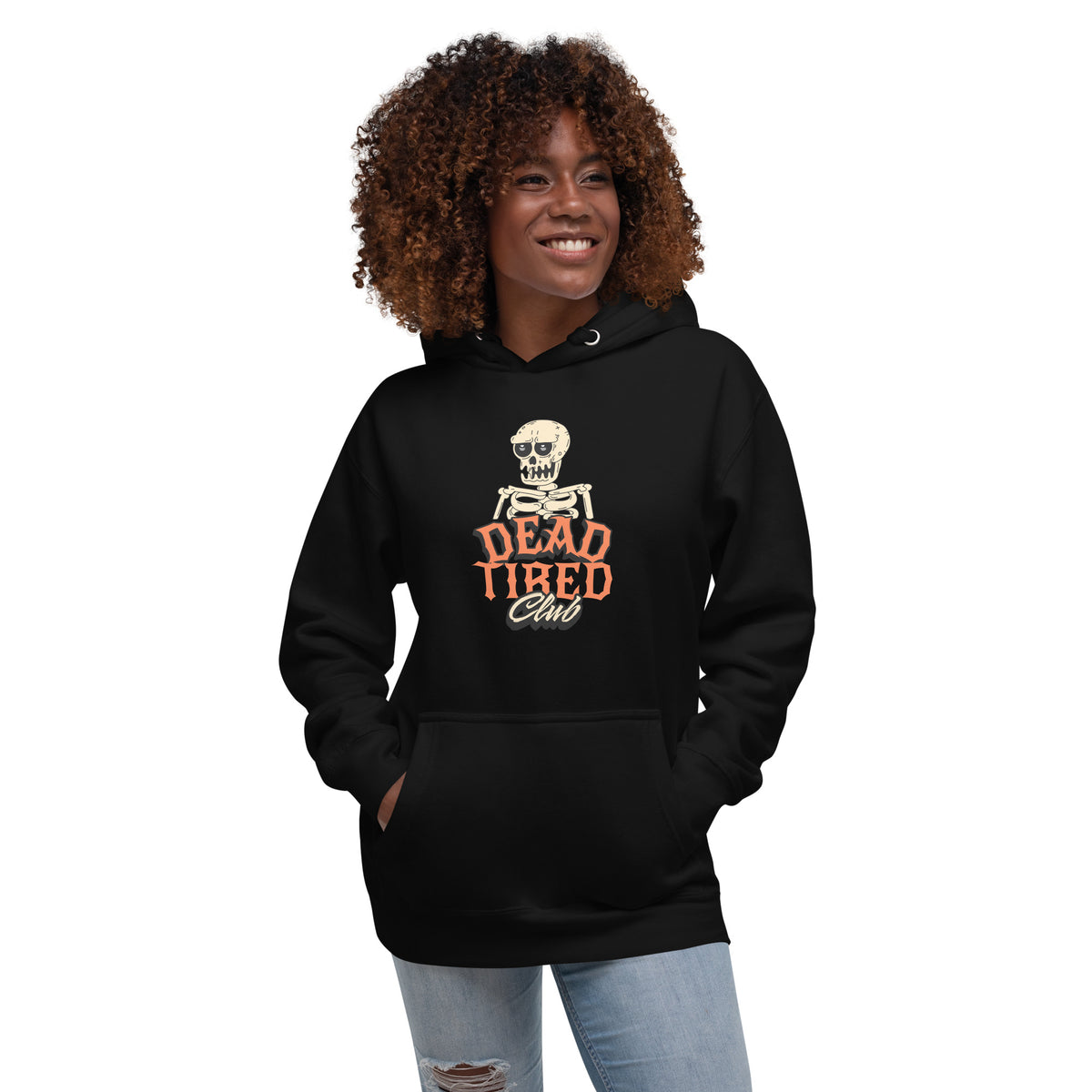 A woman with curly brown hair wearing a black pullover hoodie with a graphic print of a skeleton and the text 'DEAD TIRED Club' in orange and white. She is also wearing light blue ripped jeans, set against a plain white background.