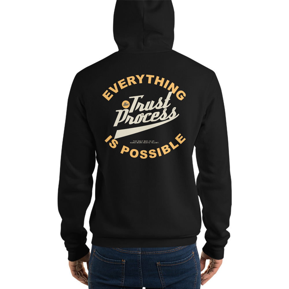 A person shown from the back wearing a black hoodie with a large cream and gold graphic on the back that reads “Everything Trust Process Is Possible,” paired with blue jeans against a plain white background.