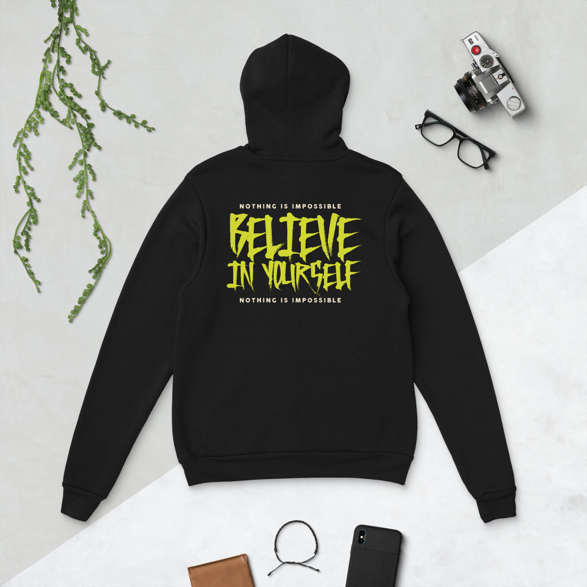 A black hoodie laid flat on a light surface, shown from the back with a large yellow graphic text reading 'BELIEVE IN YOURSELF' and smaller white text above and below. Styled flat-lay props include a plant, camera, glasses, phone, and wallet.