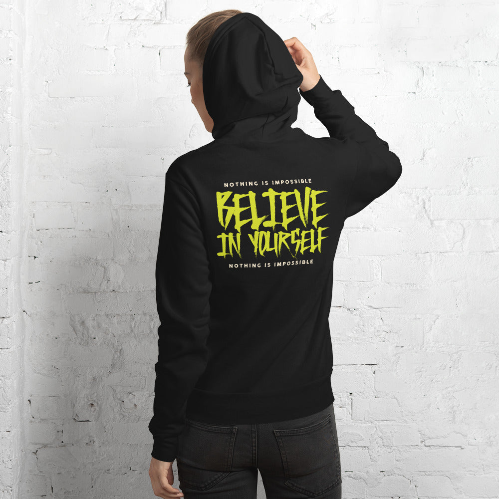 A person shown from the back wearing a black hoodie with a large yellow graphic text print reading 'BELIEVE IN YOURSELF' and smaller white text above and below, standing against a white brick wall.