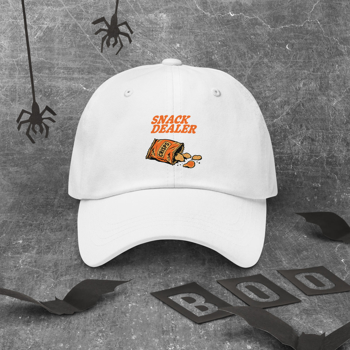 A white baseball cap with an orange 'SNACK DEALER' graphic and a candy bag illustration on the front, displayed on a textured gray background with Halloween-themed black spider and bat decorations.