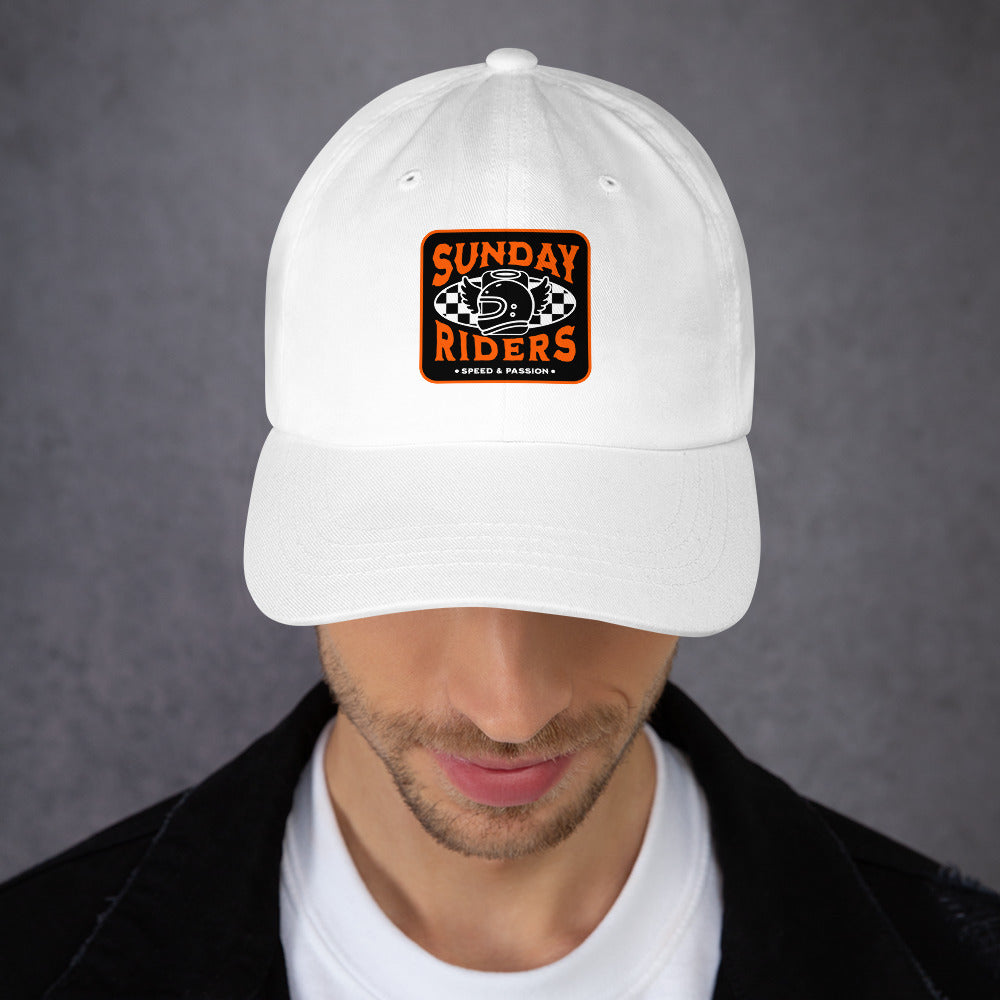 A man wearing a white baseball cap with an orange and black 'Sunday Riders' patch on the front, photographed against a textured gray background.