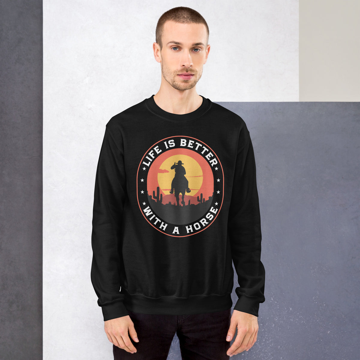 A man wearing a black crewneck sweatshirt with a circular graphic on the chest that reads 'LIFE IS BETTER WITH A HORSE' around a silhouette of a cowboy against a sunset.