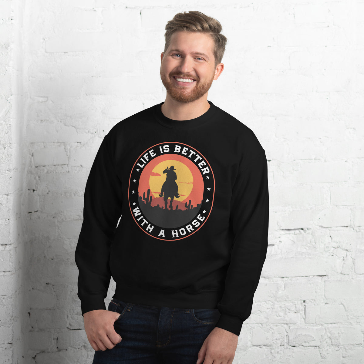 A smiling man wearing a black crewneck sweatshirt with a circular graphic that reads 'LIFE IS BETTER WITH A HORSE' around a cowboy silhouette, standing against a white brick wall.