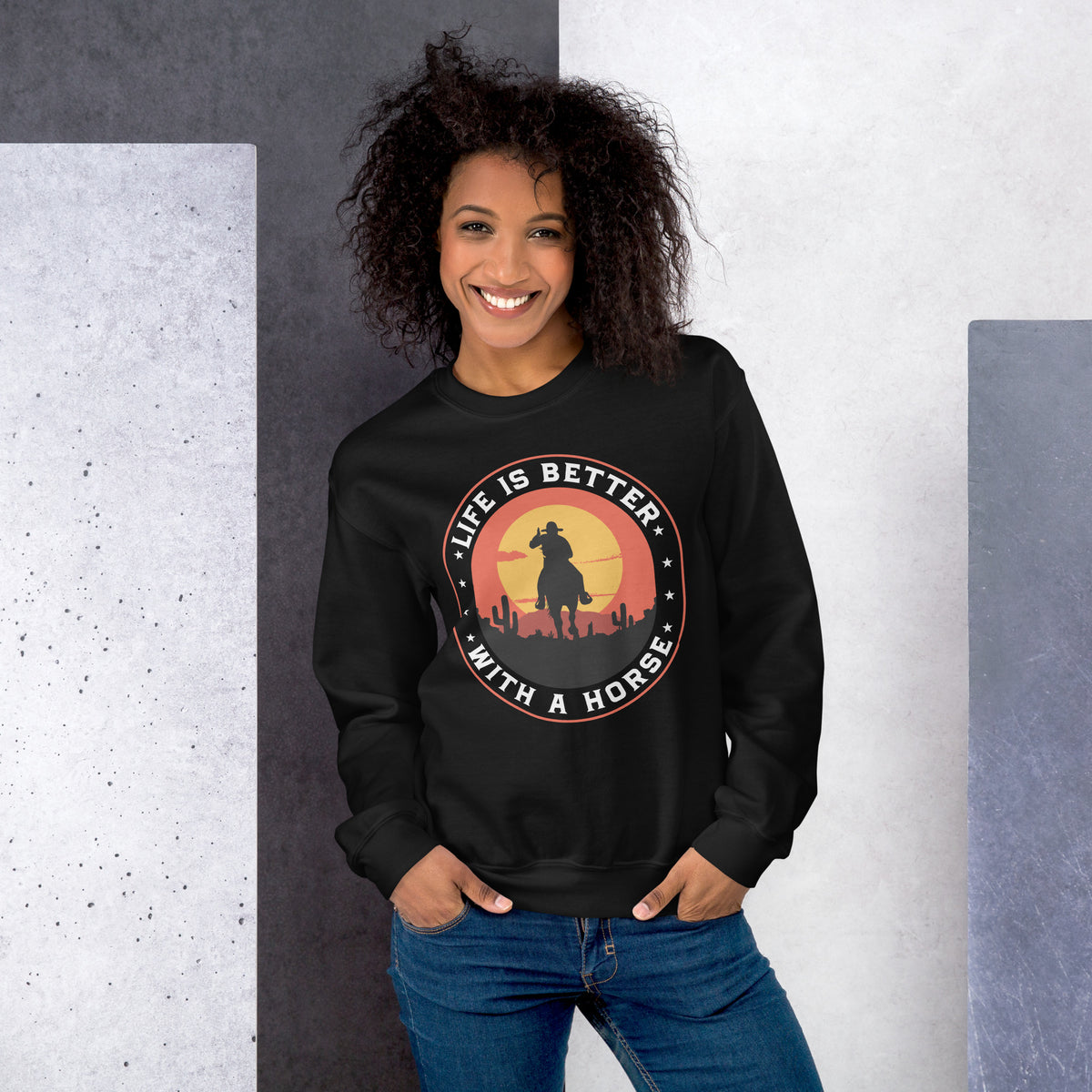A smiling woman with curly hair wearing a black crewneck sweatshirt with a circular graphic that reads 'LIFE IS BETTER WITH A HORSE' around a sunset silhouette, paired with blue jeans, standing against a split gray and white textured background.