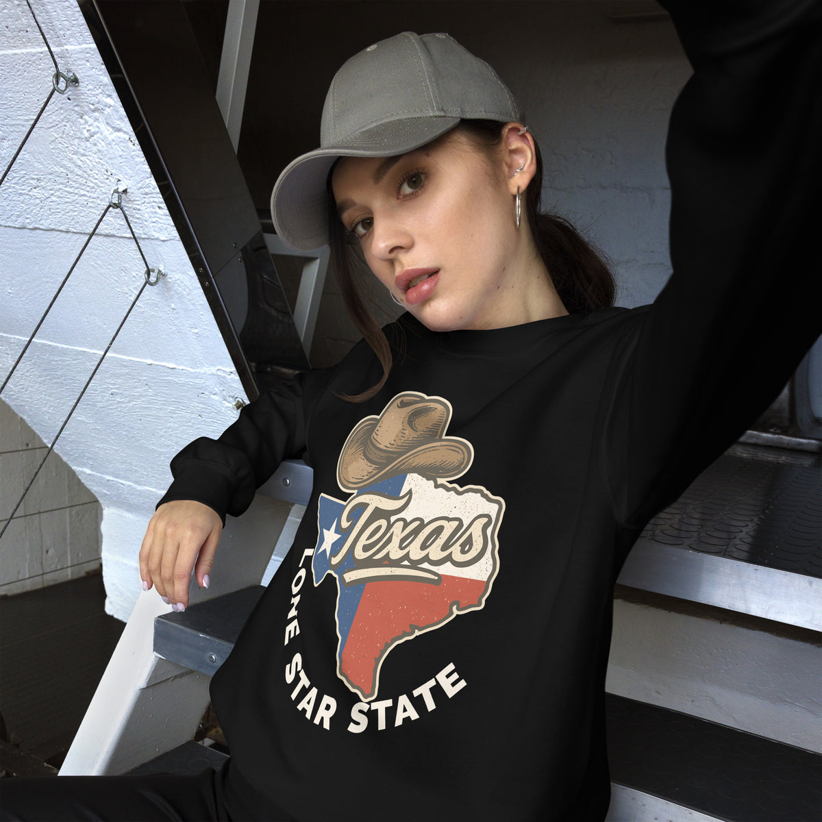 A woman wearing a black long-sleeve crewneck sweatshirt with a 'Texas Star State' graphic on the front, paired with a gray baseball cap, sitting on outdoor stairs.