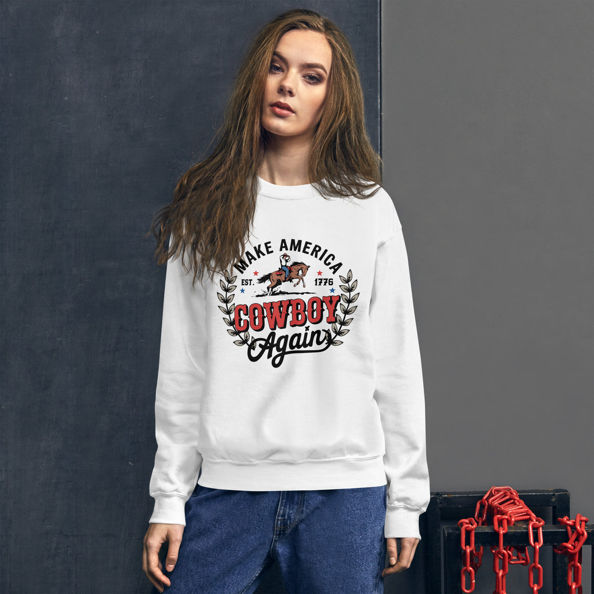 A woman wearing a white crewneck sweatshirt with a vintage-style 'Make America Cowboy Again' graphic on the chest, paired with blue jeans, standing against a dark gray textured wall.