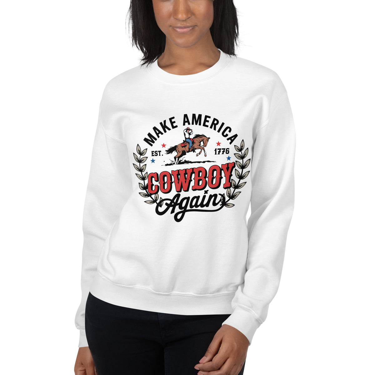 A woman wearing a white crewneck sweatshirt with a graphic print that reads 'MAKE AMERICA COWBOY Again' in black and red lettering, featuring an illustration of a cowboy on a horse and a laurel wreath. She is also wearing black pants against a plain white background.