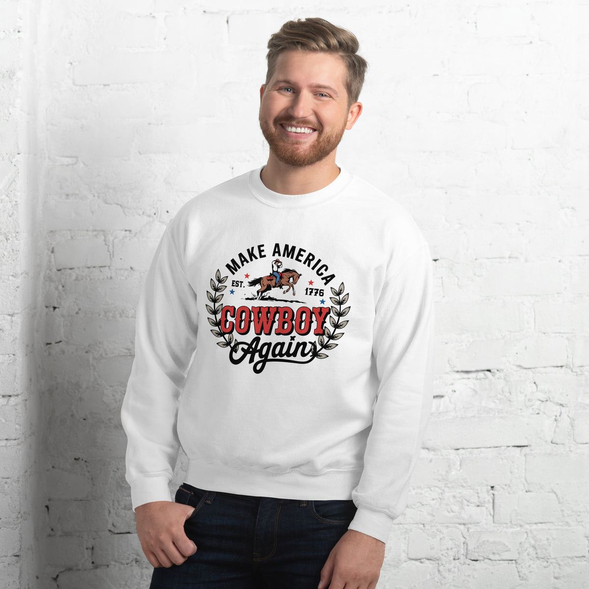 A smiling man wearing a white crewneck sweatshirt with a western-themed graphic that reads 'MAKE AMERICA COWBOY Again' in black and red lettering, posed against a white brick wall.