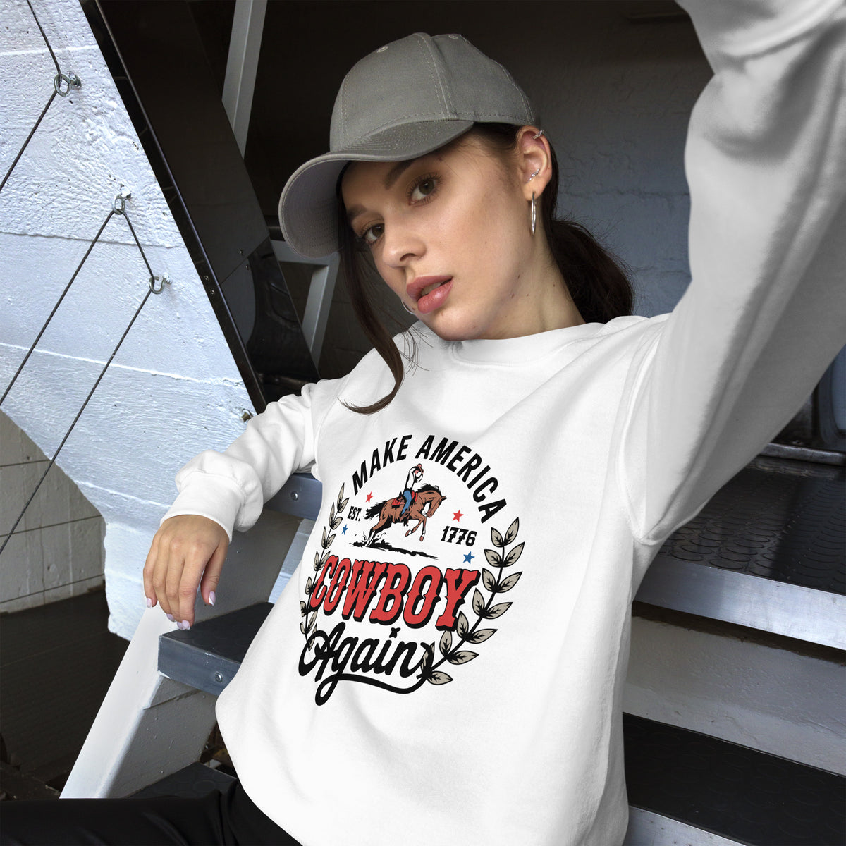 A woman wearing a white long-sleeve graphic sweatshirt with a 'Cowboy Again' and 'Make America 1776' print, paired with a gray baseball cap, sitting on outdoor steps.
