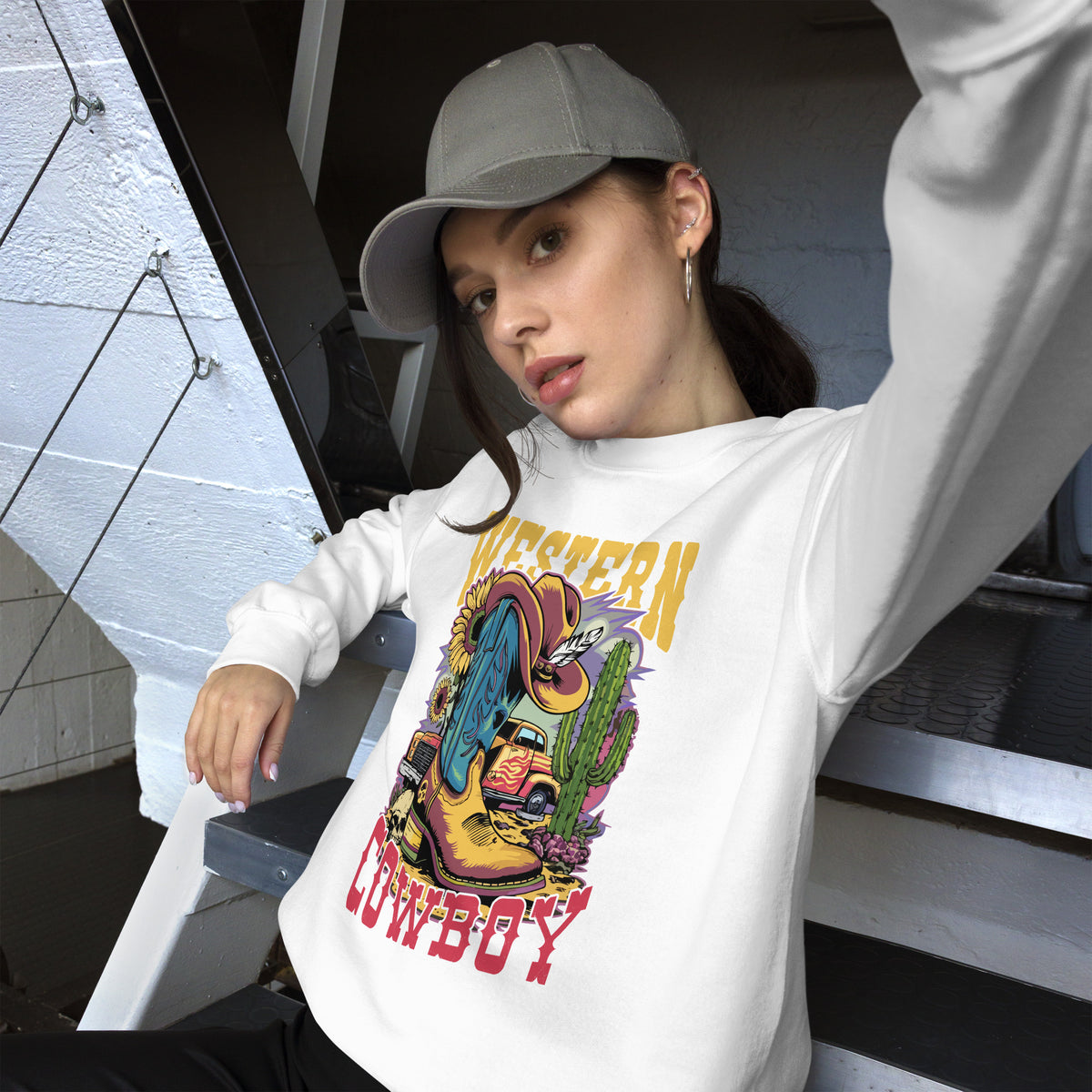A woman wearing a white long-sleeve crewneck sweatshirt with a colorful cartoon cowboy boot graphic and 'Western Cowboy' text, paired with a gray baseball cap, posing on outdoor stairs.