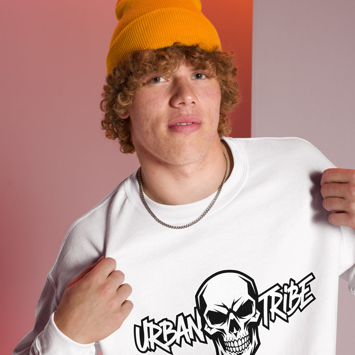 A young man with curly hair wearing a bright yellow beanie and a white crewneck sweatshirt with a black skull graphic and the text 'URBAN TRIBE'. He is posing against a split pink and orange background.