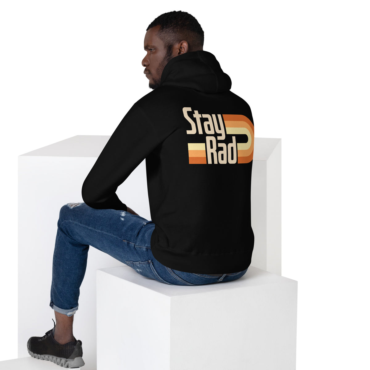 A man wearing a black hoodie with a retro-style 'Stay Rad' graphic on the back, sitting on white display blocks against a white background.