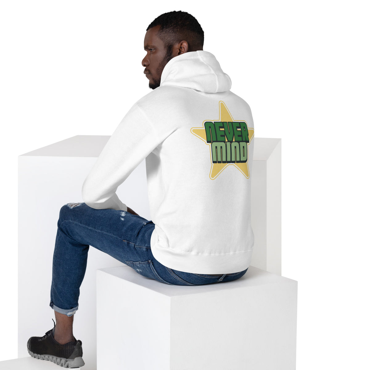 A man wearing a white hoodie with a green and yellow 'NEVER MIND' star graphic on the back, sitting on white display blocks against a white background.