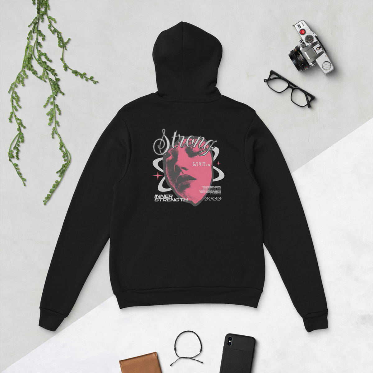A black hoodie shown from the back in a styled flat-lay, featuring a pink and white graphic with the word 'Strong' and a face illustration. The hoodie is arranged on a light surface with a camera, glasses, a phone, a wallet, and a plant sprig as props.
