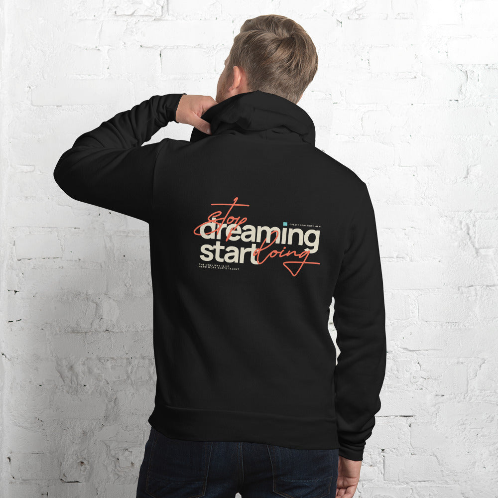 A man viewed from behind wearing a black pullover hoodie with a motivational graphic on the back that reads 'Stop dreaming and start doing' in white and orange lettering, posed against a white brick wall.