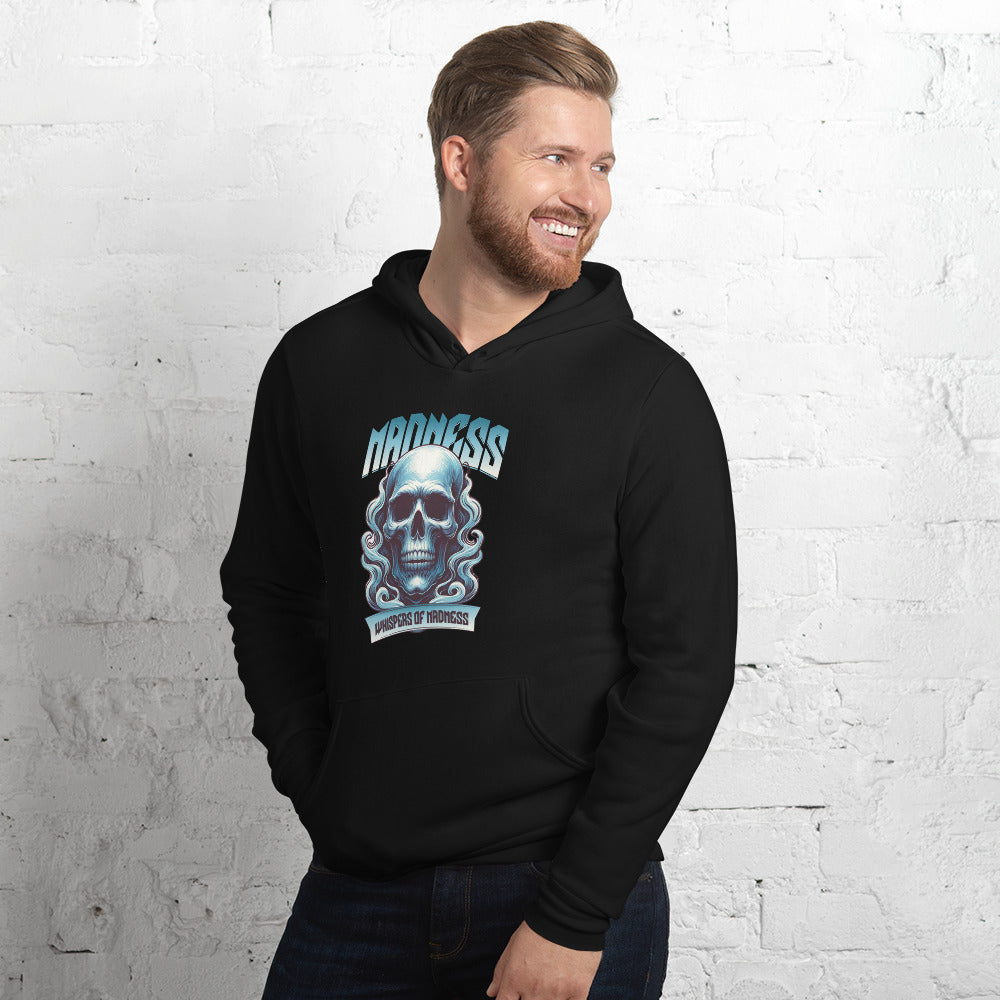 A smiling man with a beard wearing a black hoodie with a blue and white skull graphic and the word 'Madness' on the front, standing against a white brick wall.