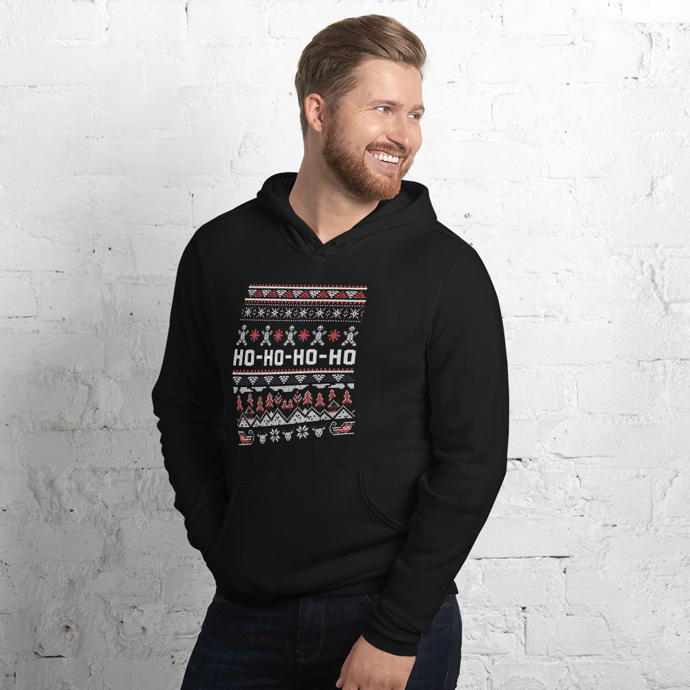 A smiling man wearing a black pullover hoodie with a festive Christmas-themed graphic print on the chest, posed against a white brick wall.
