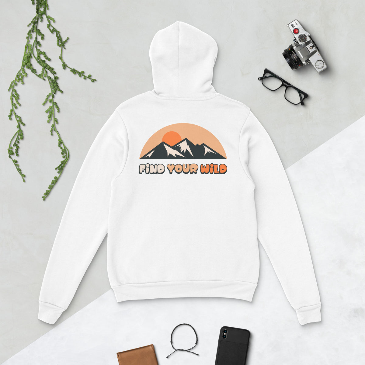 A white pullover hoodie laid flat on a light gray surface, shown from the back with a mountain-and-sun graphic and the words 'FIND YOUR WILD' in orange and black. The hoodie is styled with a camera, glasses, a phone, a wallet, and a hanging green branch around it.