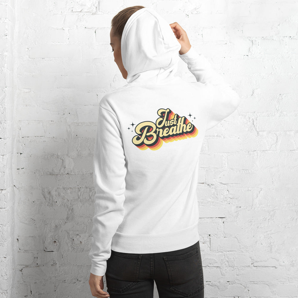 A person wearing a white hoodie with a colorful retro-style 'Just Breathe' graphic on the back, standing against a white brick wall.