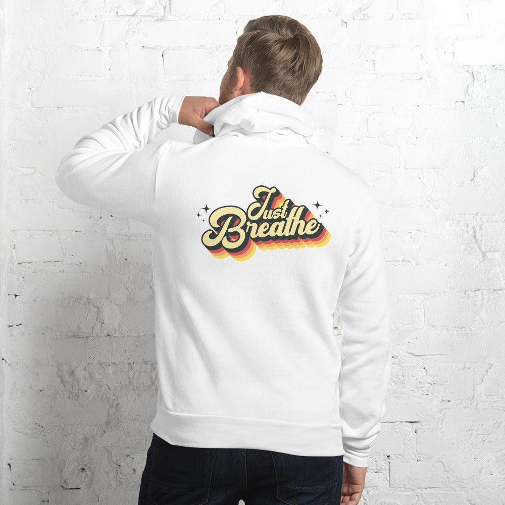 A man wearing a white hoodie with a retro-style 'Just Breathe' graphic on the back, standing against a white brick wall.