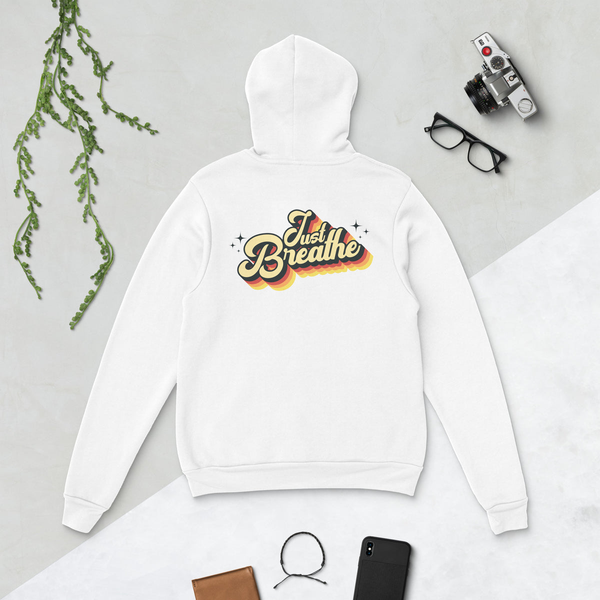 A white pullover hoodie laid flat on a light gray surface, shown from the back with a retro-style 'Just Breathe' graphic in yellow, orange, and brown. The hoodie is styled with a camera, glasses, a phone, a wallet, and a hanging green branch.